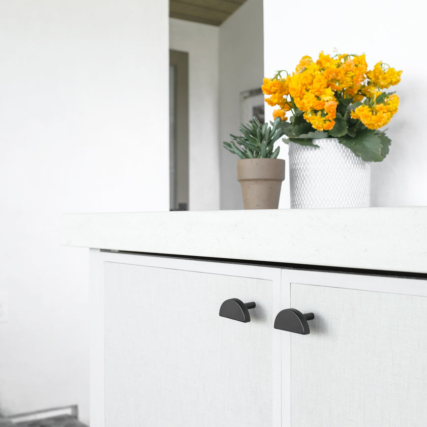 Two Black Moon knobs installed on white cabinets. There are orange flowers on the countertop.