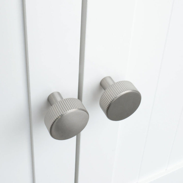 Two textured satin nickel knobs on white kitchen cabinets with ribbed exteriors. The knobs are round.